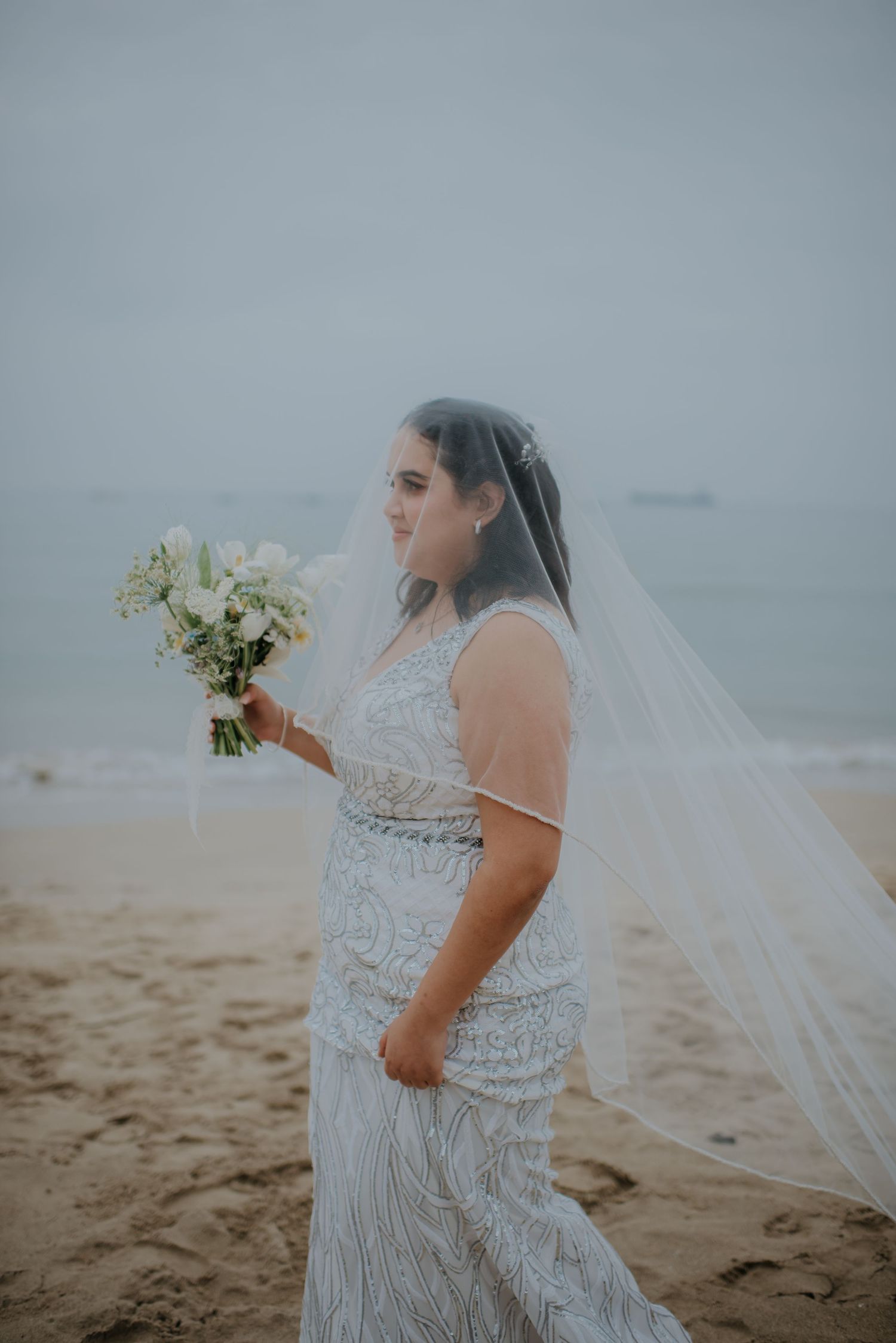 A bride in a detailed lace wedding dress holds a white bouquet while standing on a misty beach.