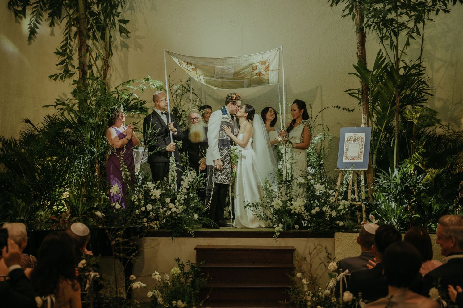 Wedding ceremony in progress on an elevated platform with floral decorations and draped fabric overhead.