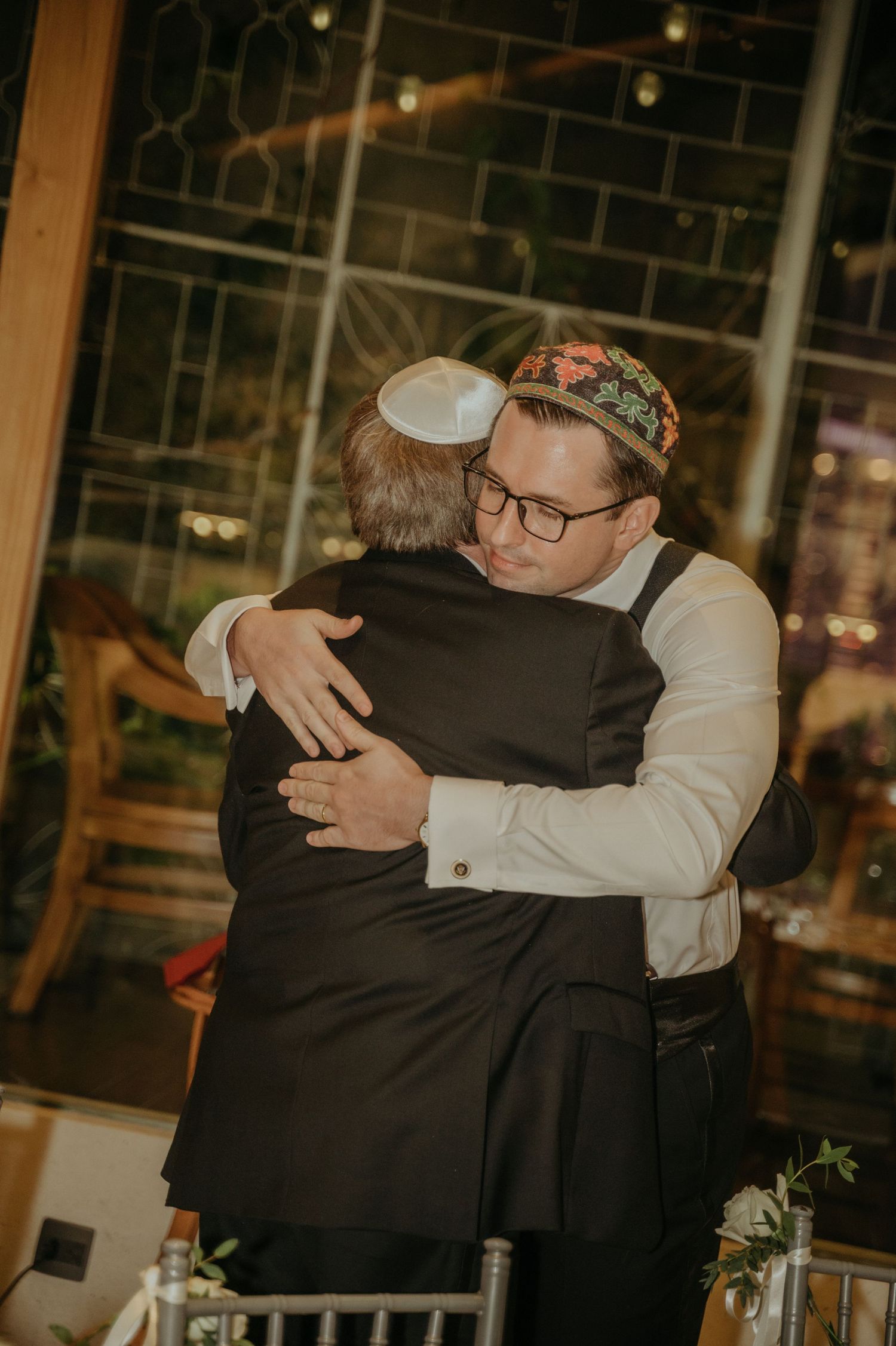 Two guests share an emotional embrace during a wedding reception in an elegantly lit venue.