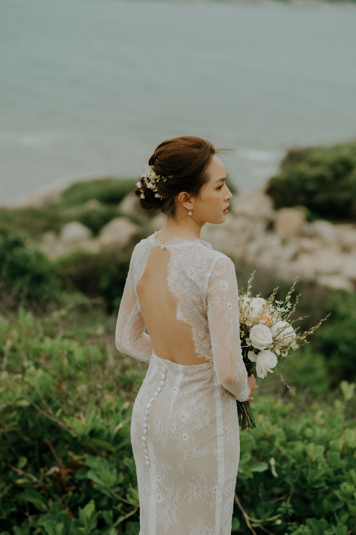 Elegant white lace wedding dress with open back design and delicate bouquet photographed against coastal scenery.