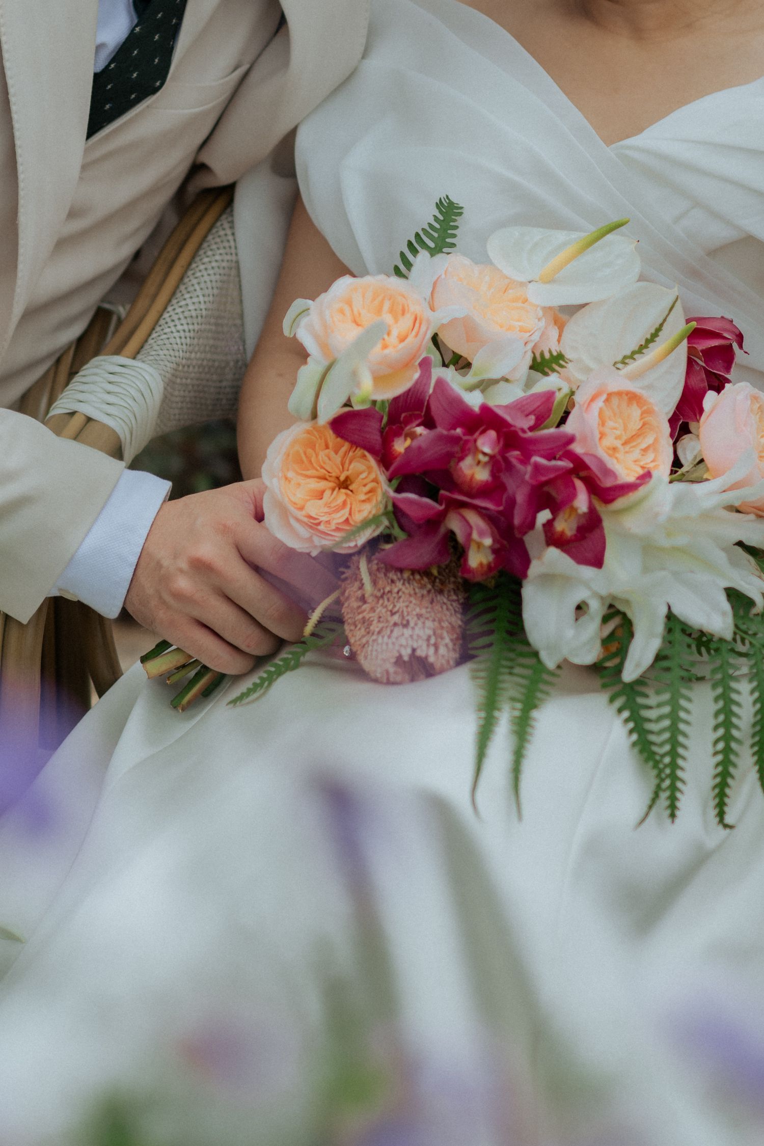 Close-up of a bridal bouquet with peach garden roses, magenta orchids, white lilies and ferns against a white dress.