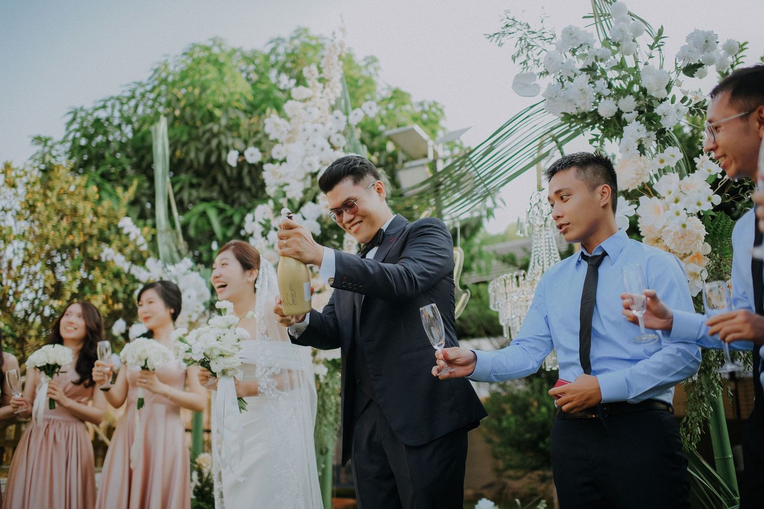 Wedding party celebrates outdoors with champagne toast among garden decorations.