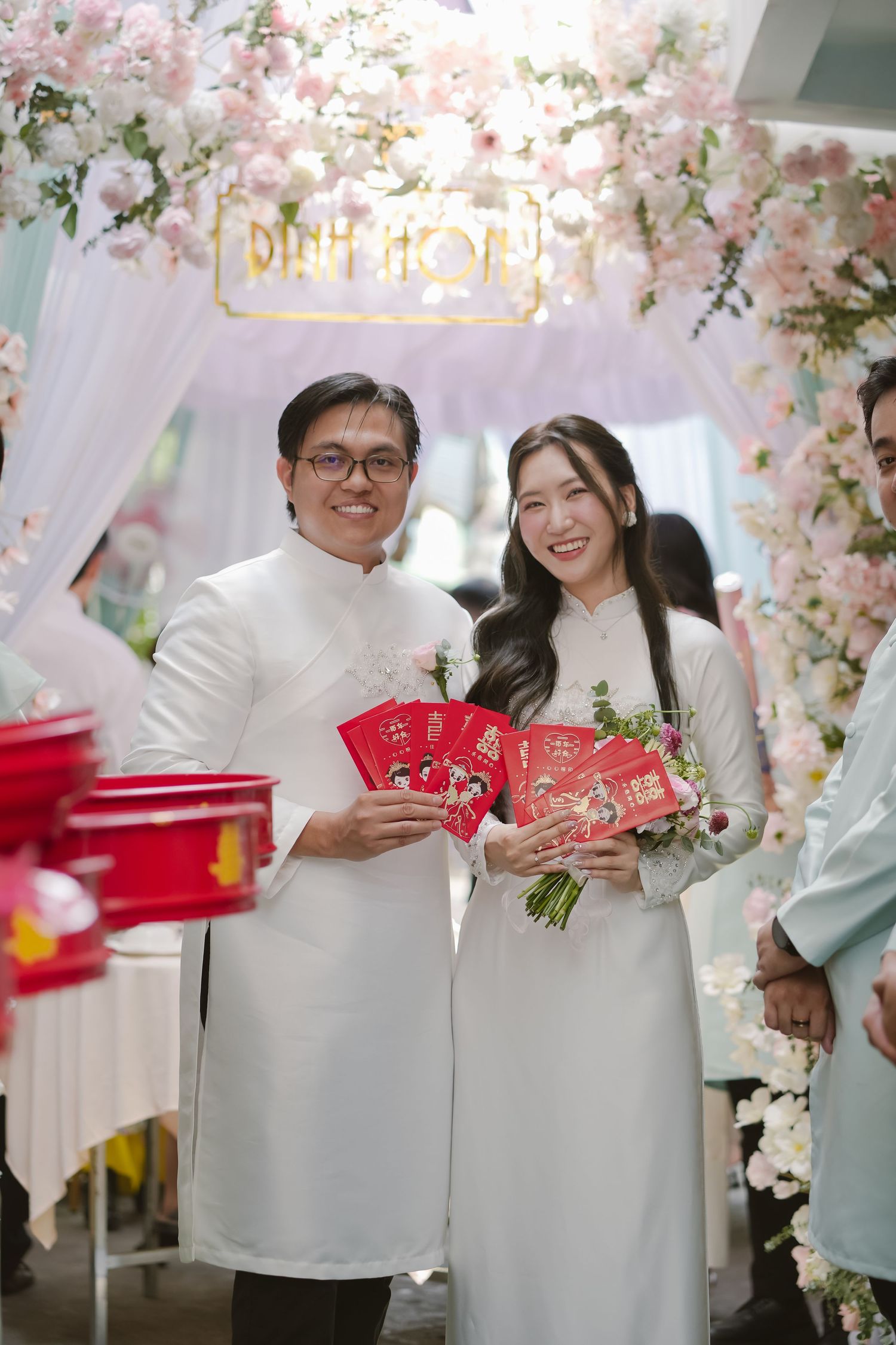 Couple in white ao dai holding red envelopes under floral arch during wedding ceremony.