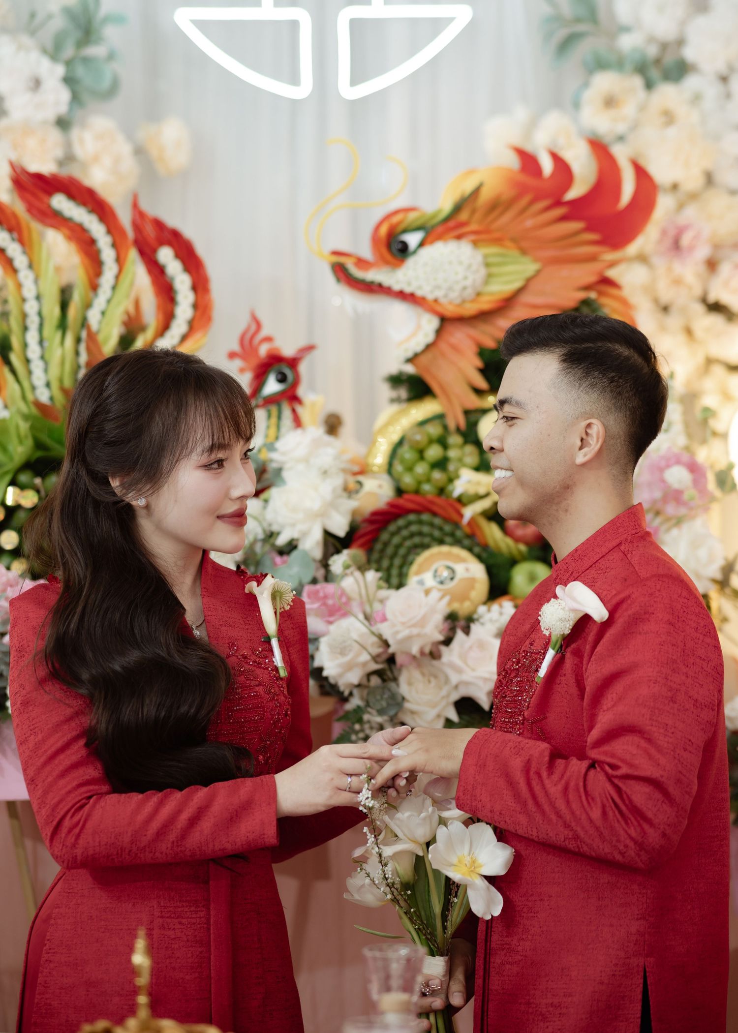A couple in matching red outfits stands together at an event with floral decorations and phoenix designs in the background.