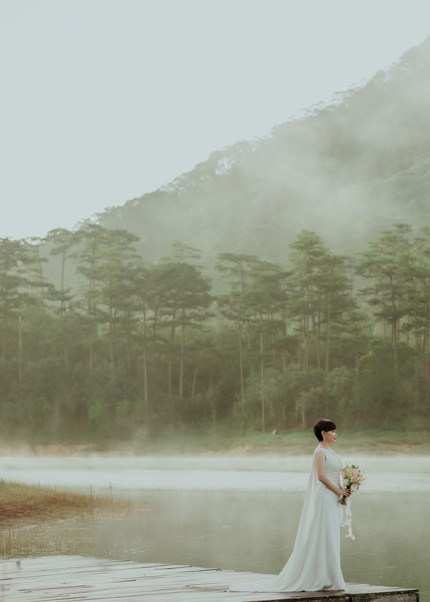 A figure in a white wedding dress stands alone by a misty lake surrounded by pine trees on a foggy morning.