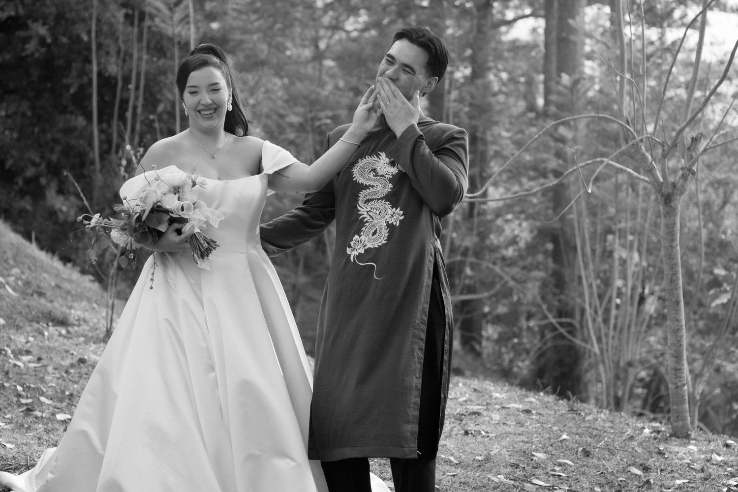 A couple in a wedding dress and long coat dance together in a black and white photograph taken outdoors.