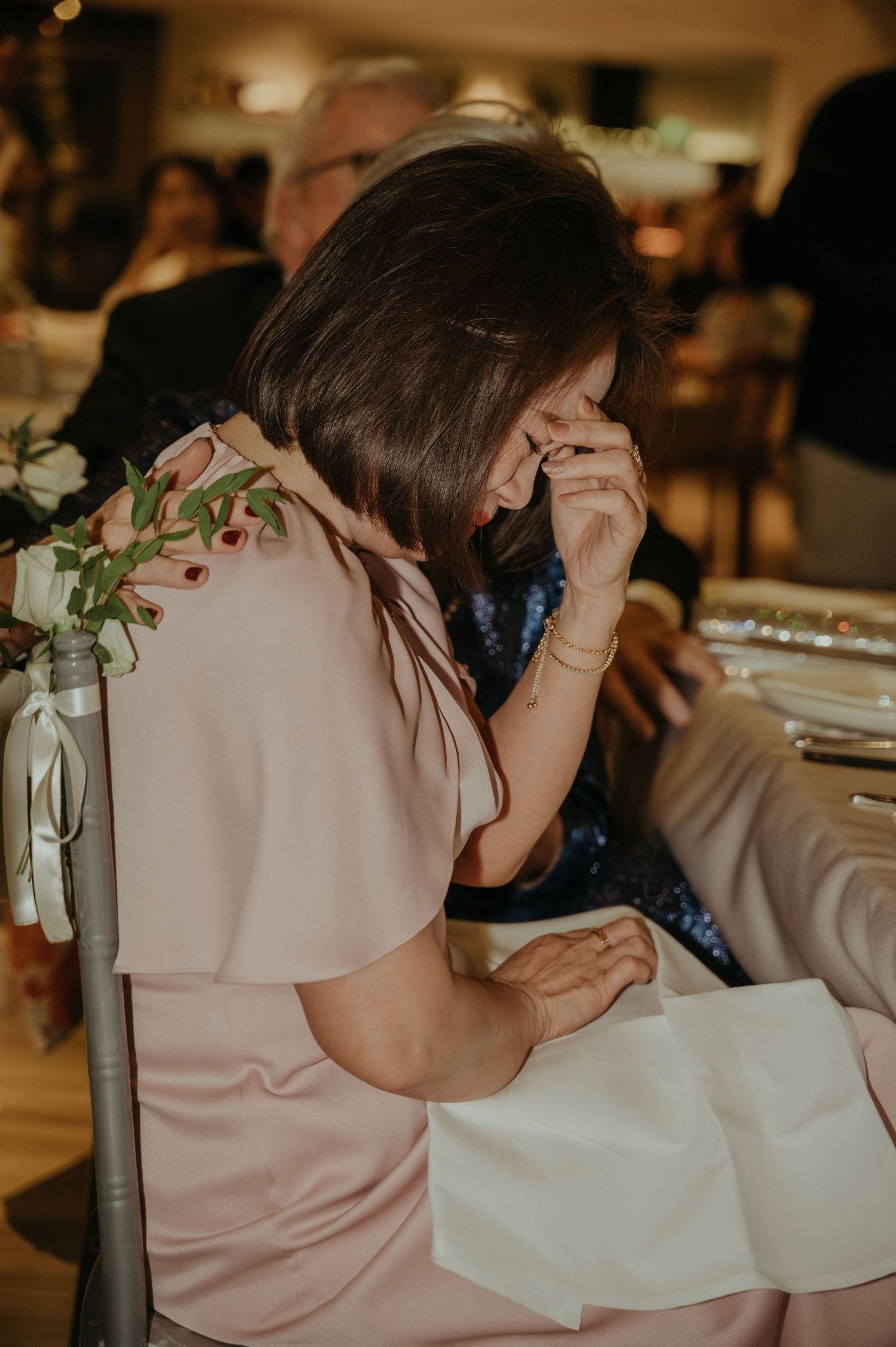 Wedding guest in pink dress wipes away tears during emotional moment at reception.