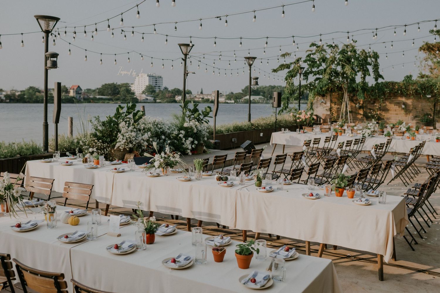 Elegant waterfront dining setup with white tablecloths and folding chairs under string lights.