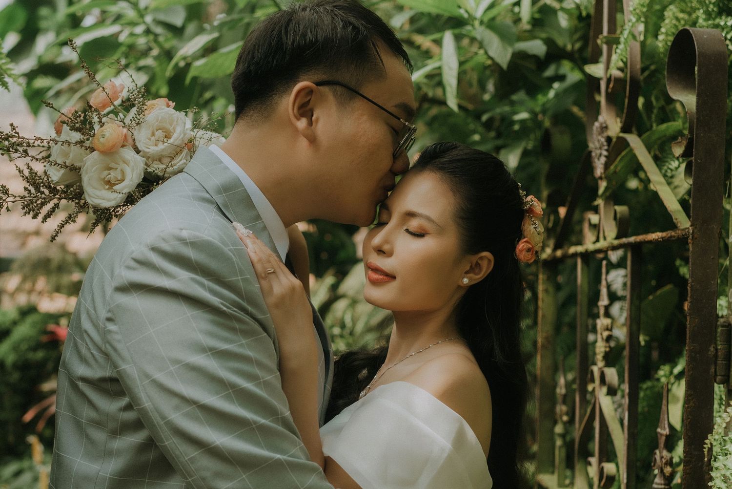 A romantic couple in wedding attire embrace in a lush garden setting with natural greenery in the background.