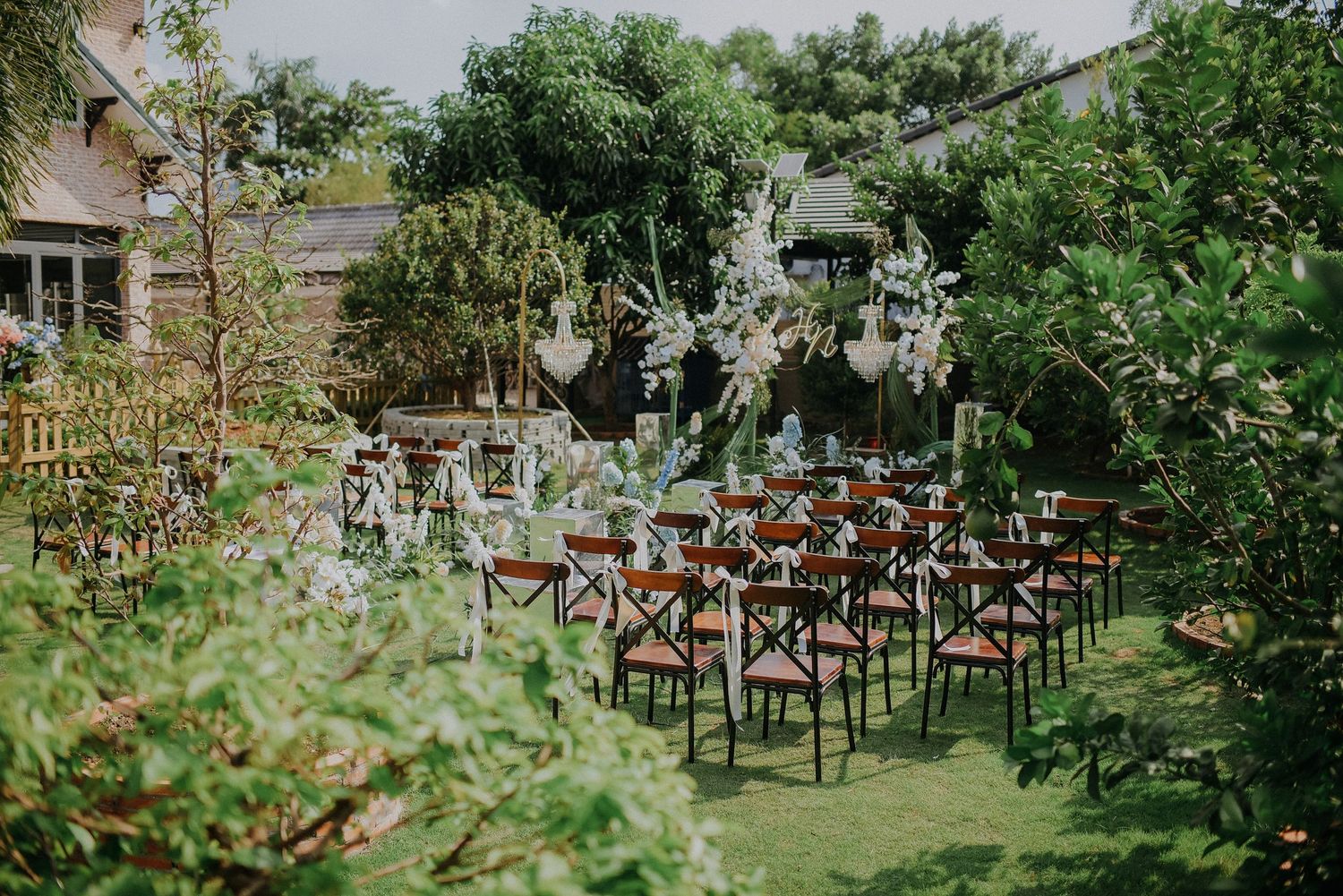 Garden wedding venue setup with wooden cross-back chairs and floral decorations under natural greenery.