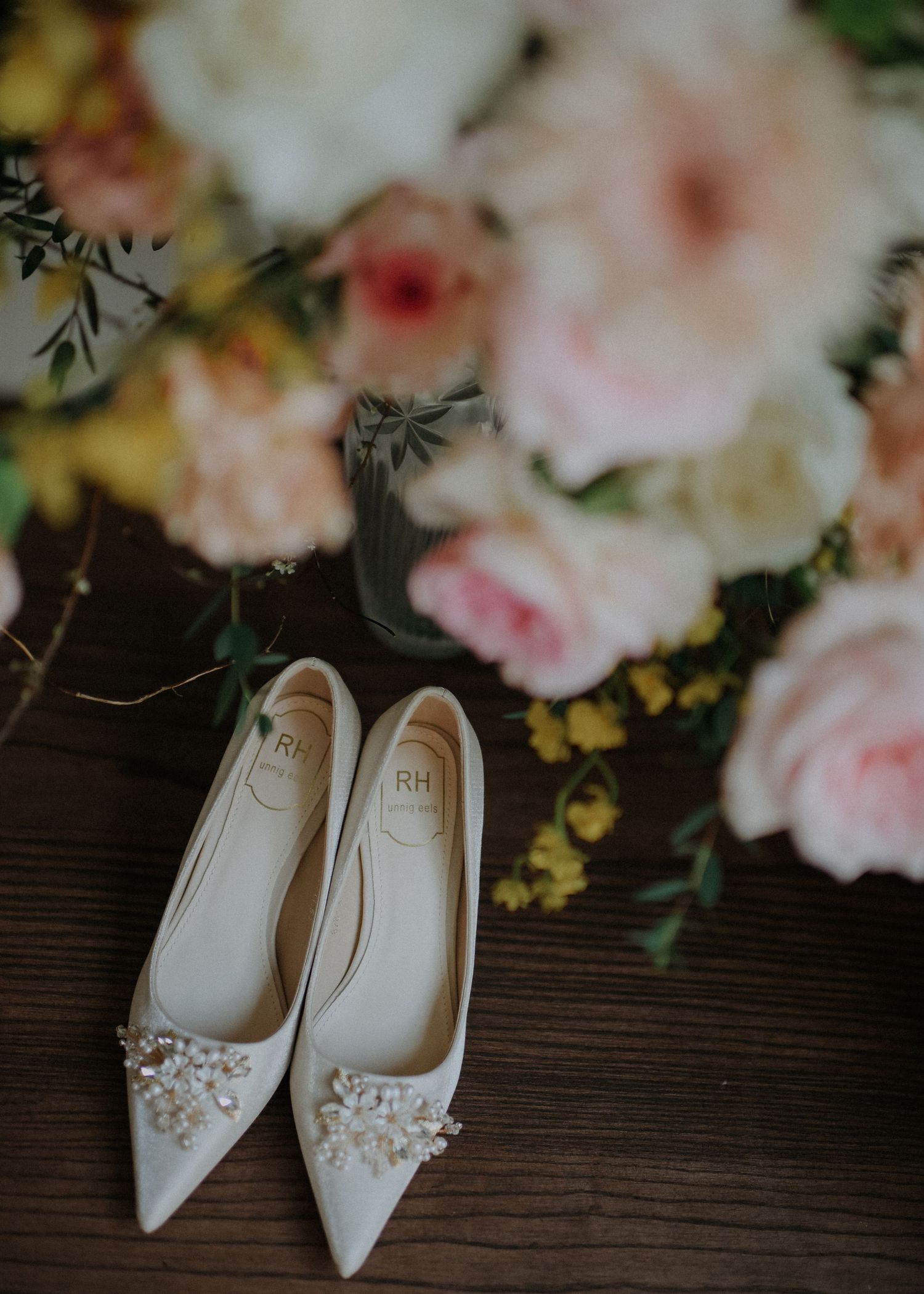 Elegant white embellished bridal shoes styled with soft pink roses on wooden surface.