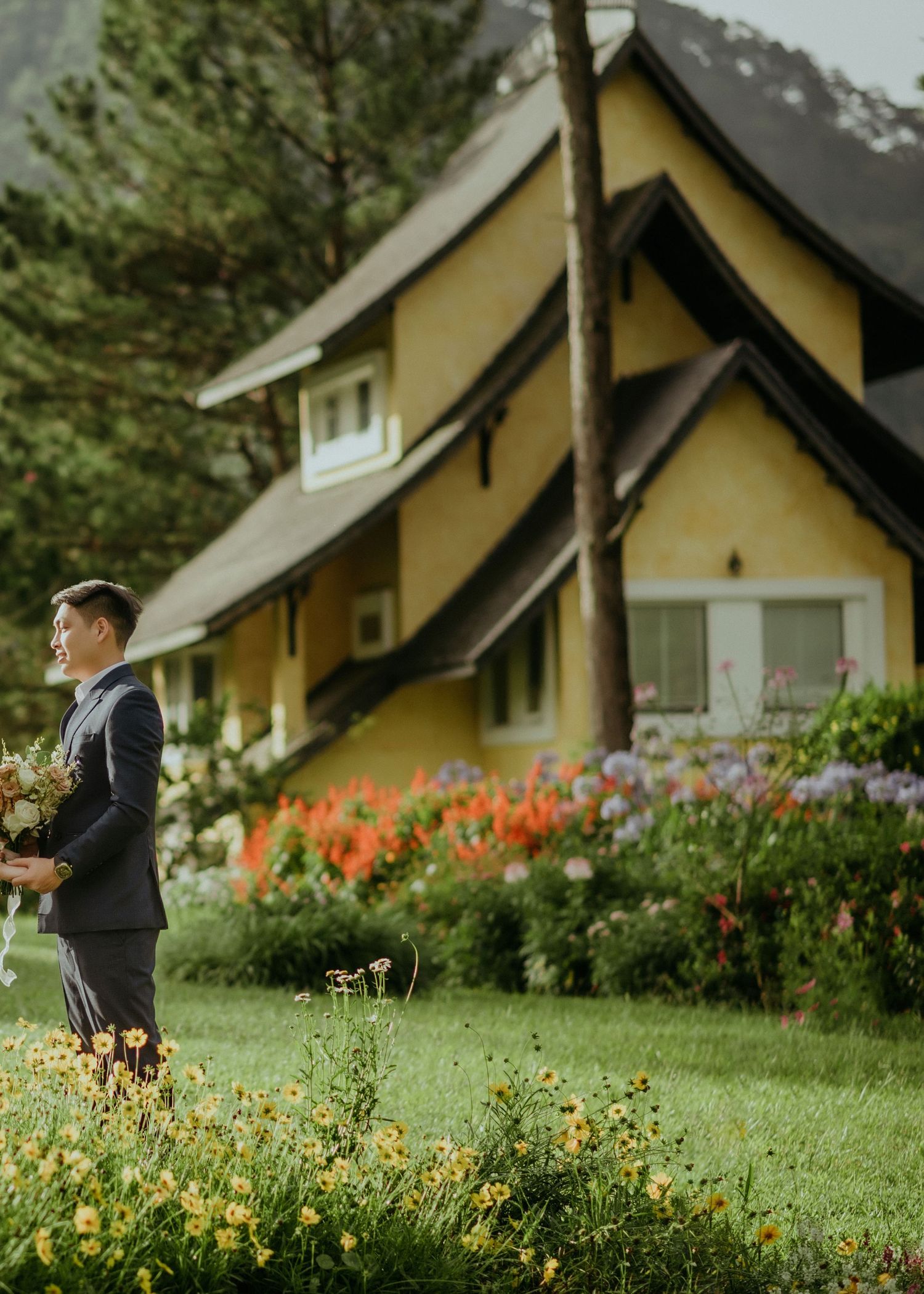 A yellow A-frame house with dark trim sits among a colorful garden with orange flowers and landscaping.