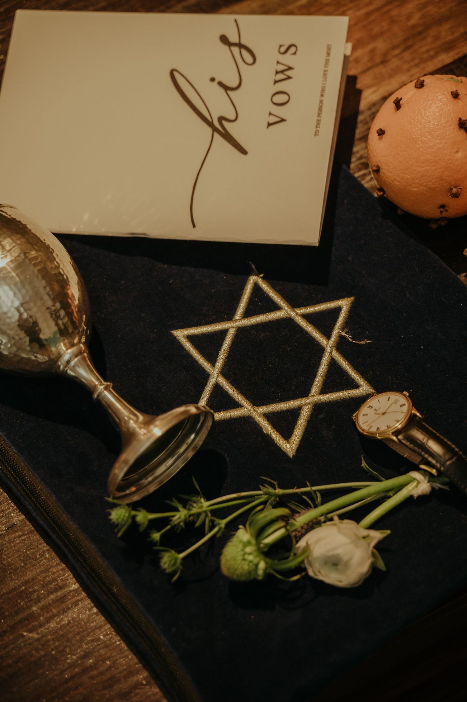 Jewish wedding details with Star of David, vows book, and ceremonial glass on dark surface.
