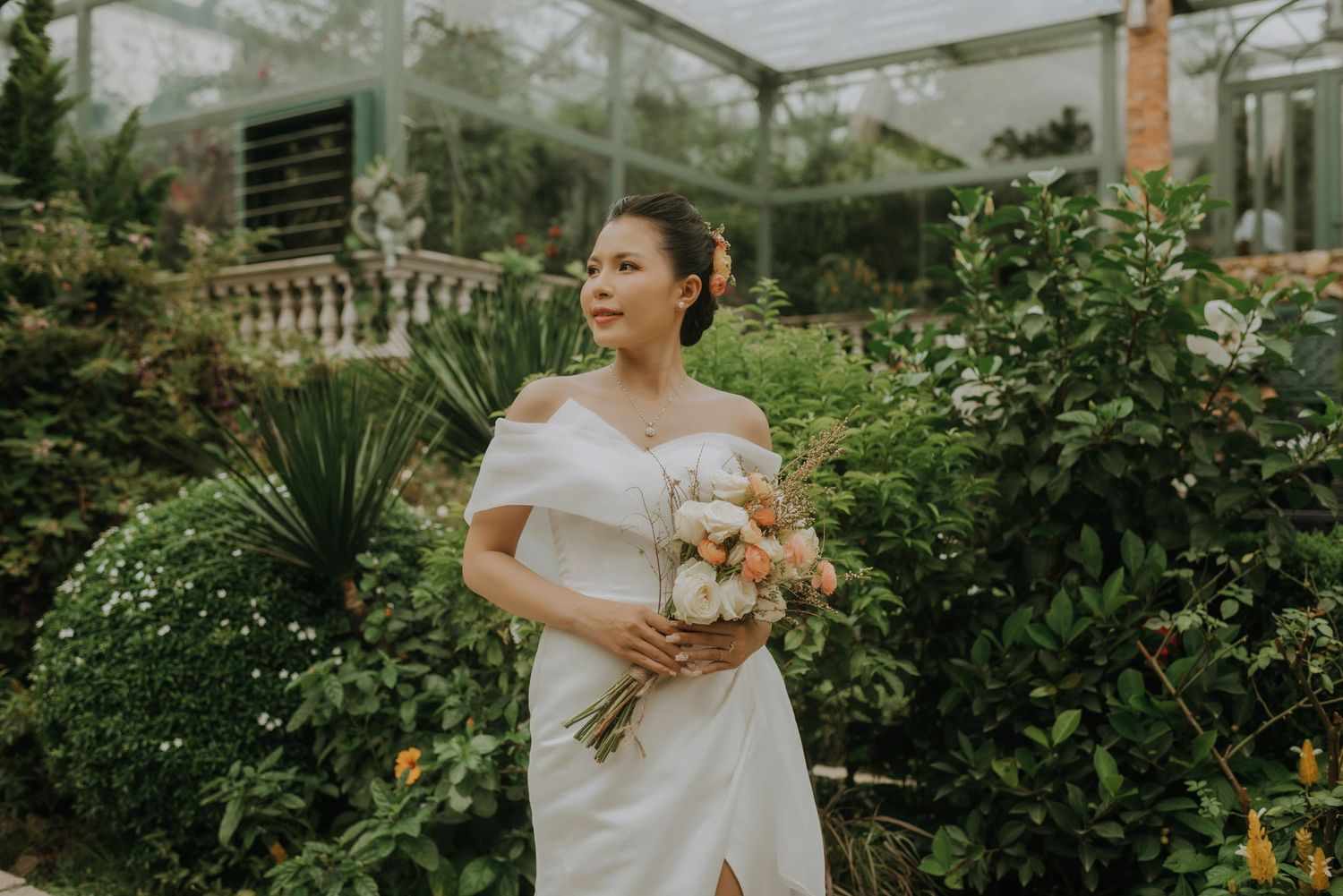 A white off-shoulder wedding dress with a soft pink bouquet in a lush greenhouse garden setting.