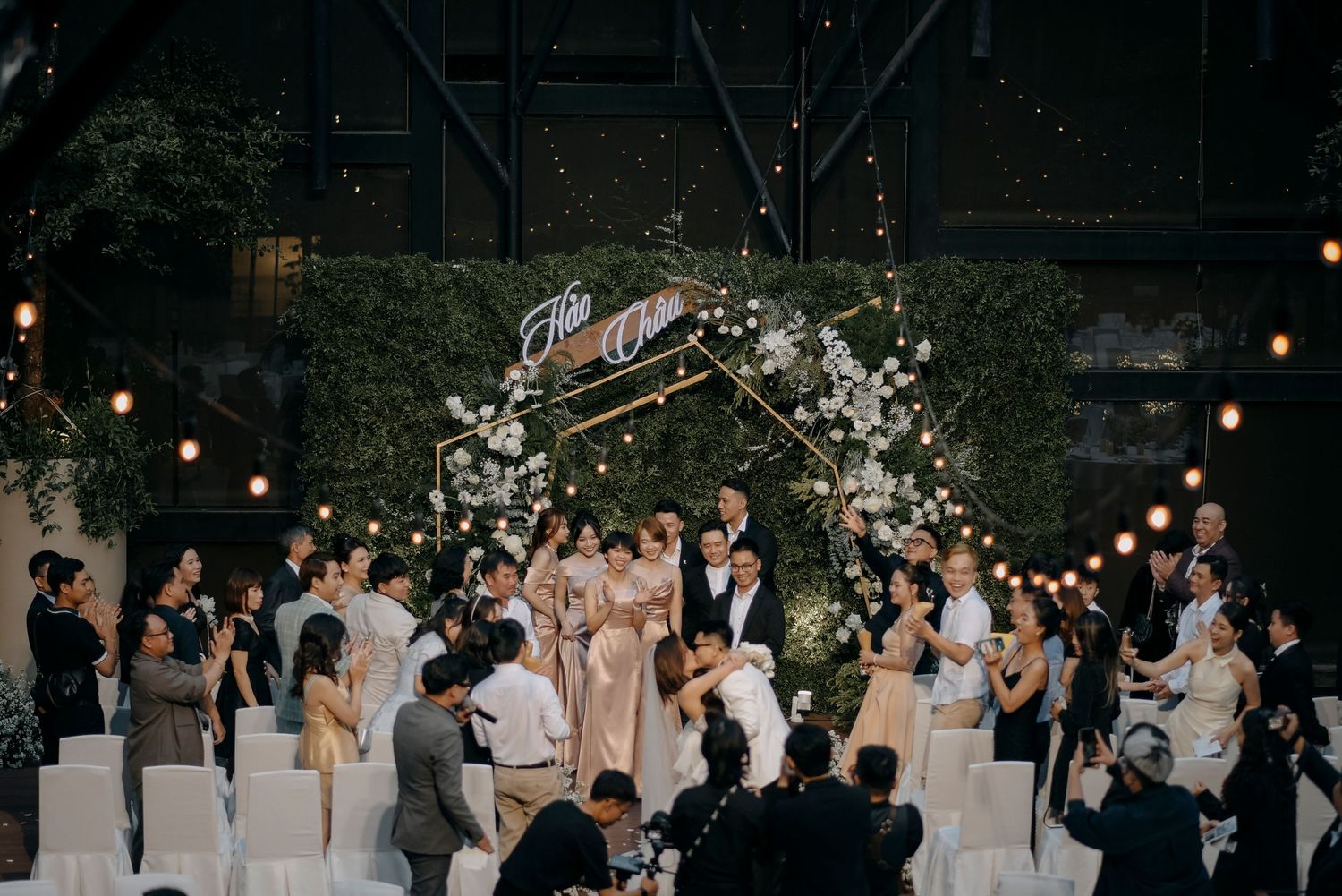 Wedding ceremony under a floral arch with guests gathered in an intimate indoor venue.
