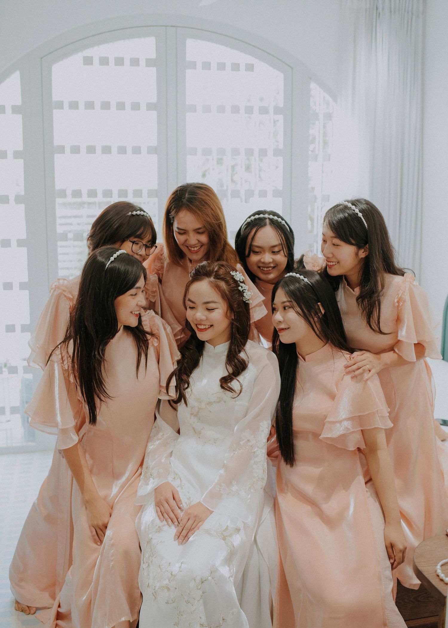 Bridesmaids in blush pink dresses gather around bride in white gown during intimate wedding moment.