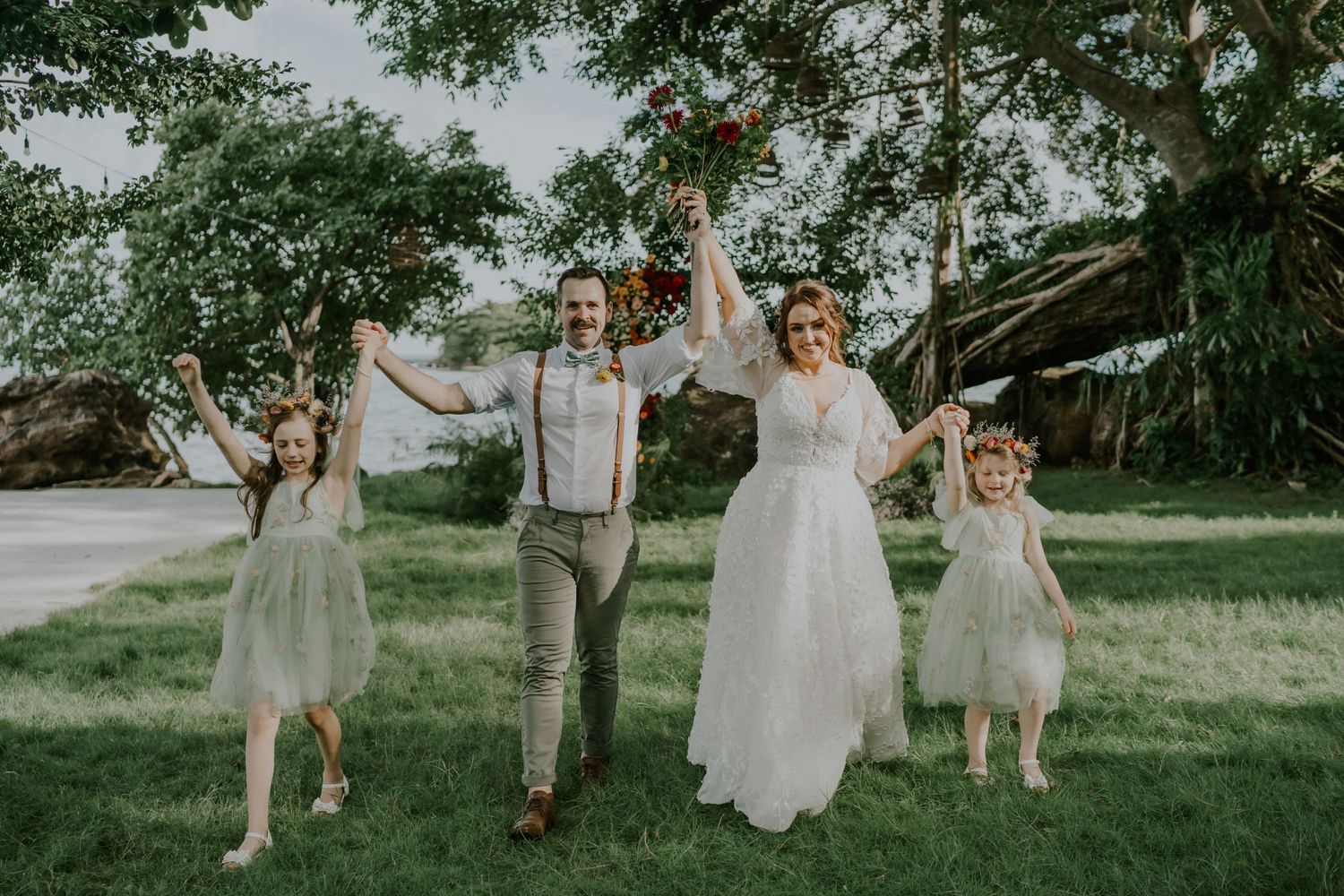 Joyful wedding party celebrates on a lush green lawn surrounded by trees with their arms raised in celebration.