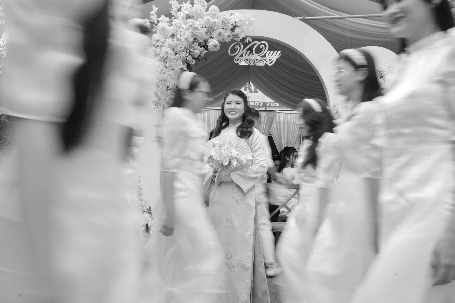 Black and white photo showing traditional wedding ceremony procession with decorative arch.
