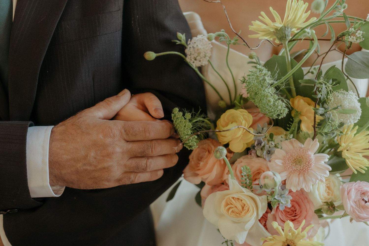 Elegant suit sleeve with formal cuff next to a pastel floral arrangement of roses, dahlias and greenery.