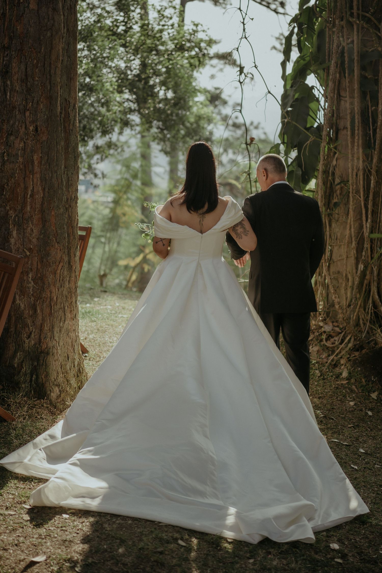 A bride wearing an off-shoulder white wedding gown and groom in black suit walking together between tall trees in a forest.