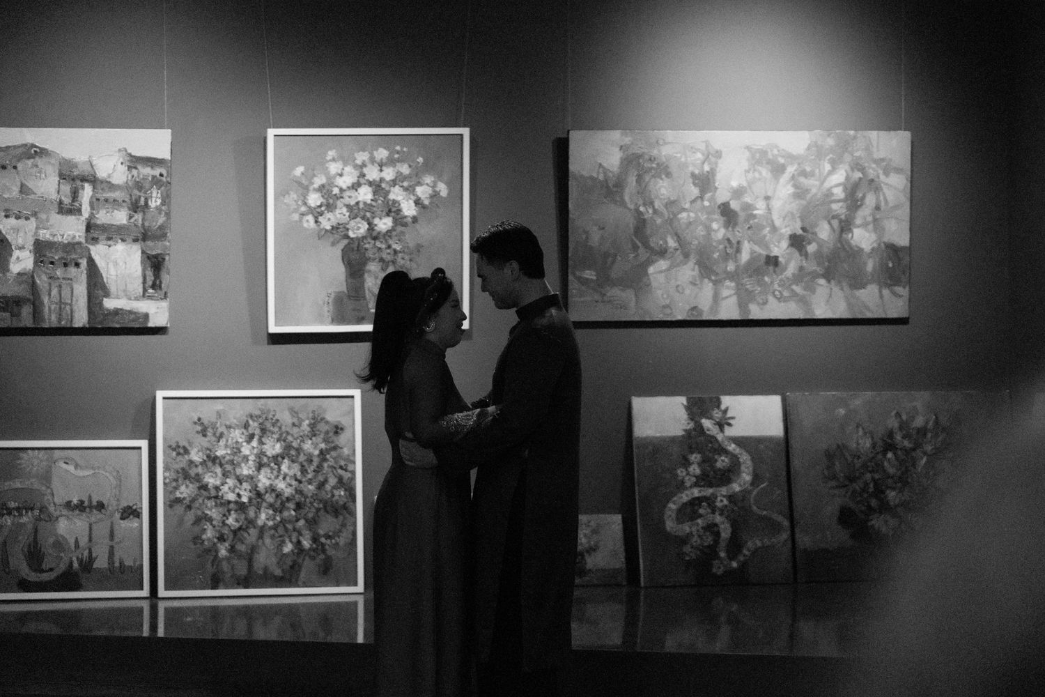 A couple stands in silhouette at an art gallery, viewing floral paintings displayed on a dark gray wall.