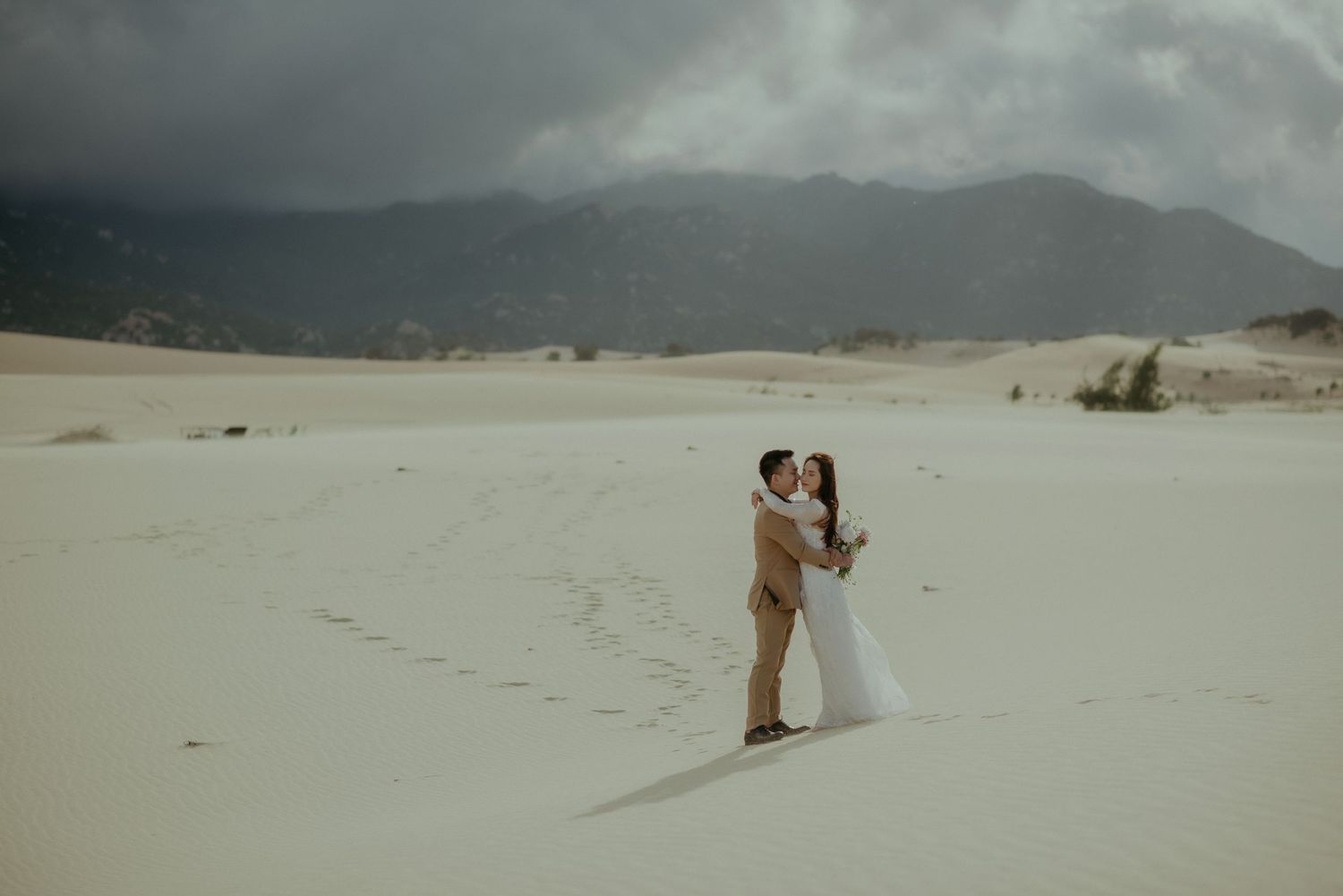 Two figures embrace in a romantic scene on sandy desert dunes with stormy mountains in the background.