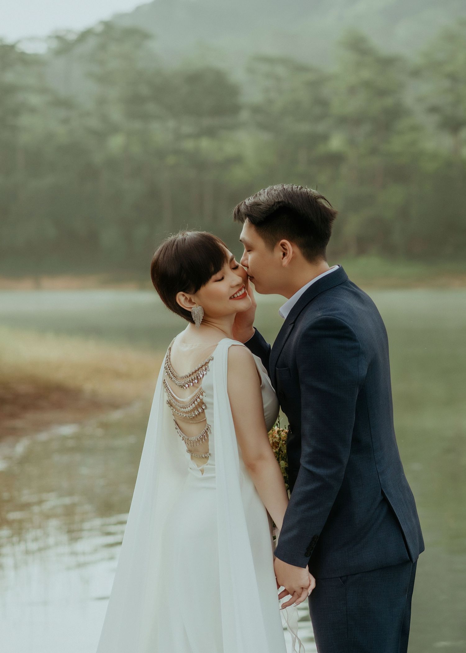A romantic couple shares a tender kiss by a misty lake, with the bride wearing an elegant backless dress with pearl details.