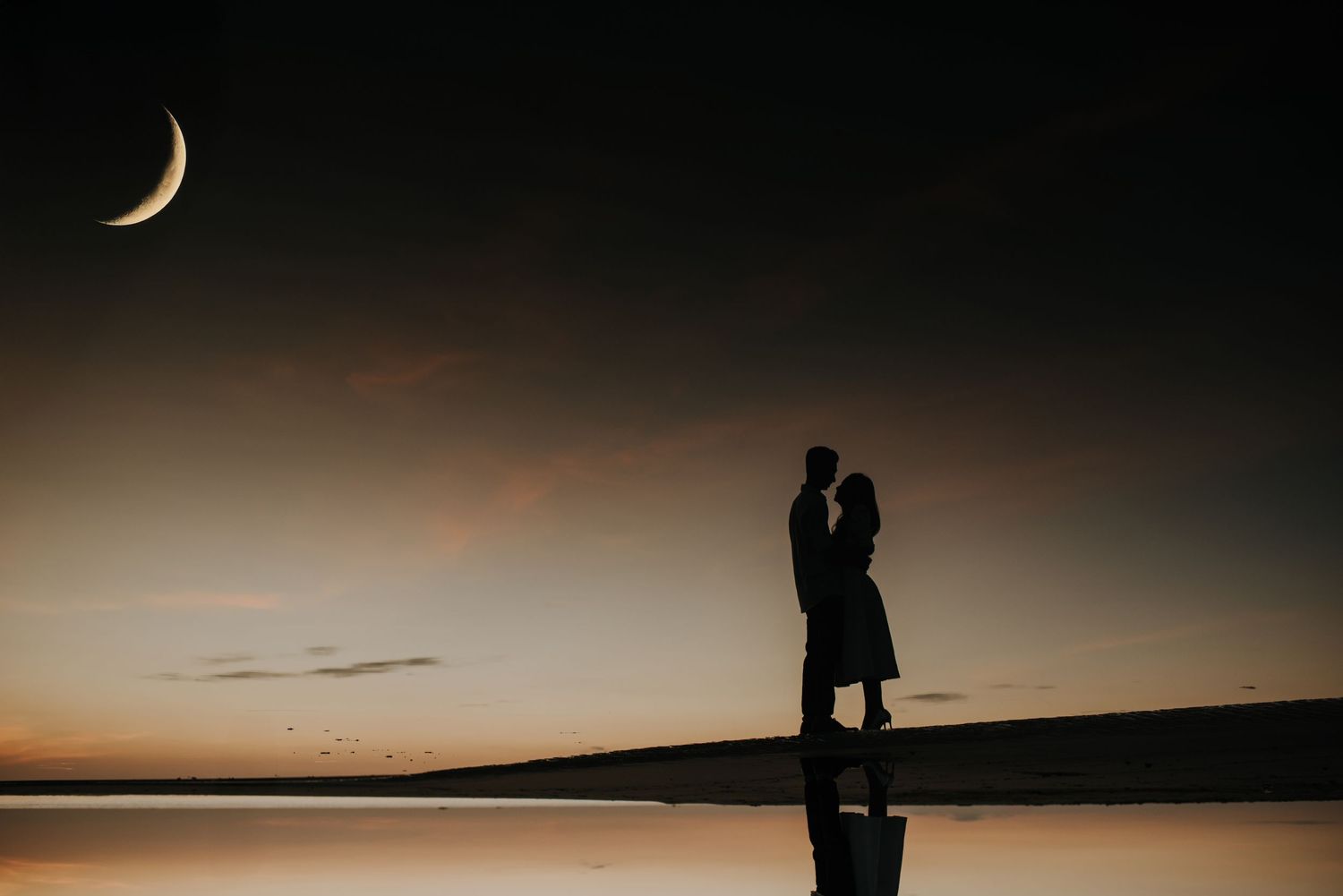 Silhouetted couple embraces on reflective surface under crescent moon during sunset.