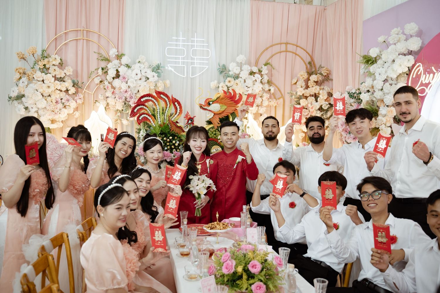 Guests at an elegant wedding reception with pink decor raise their drinks in celebration at a decorated banquet table.