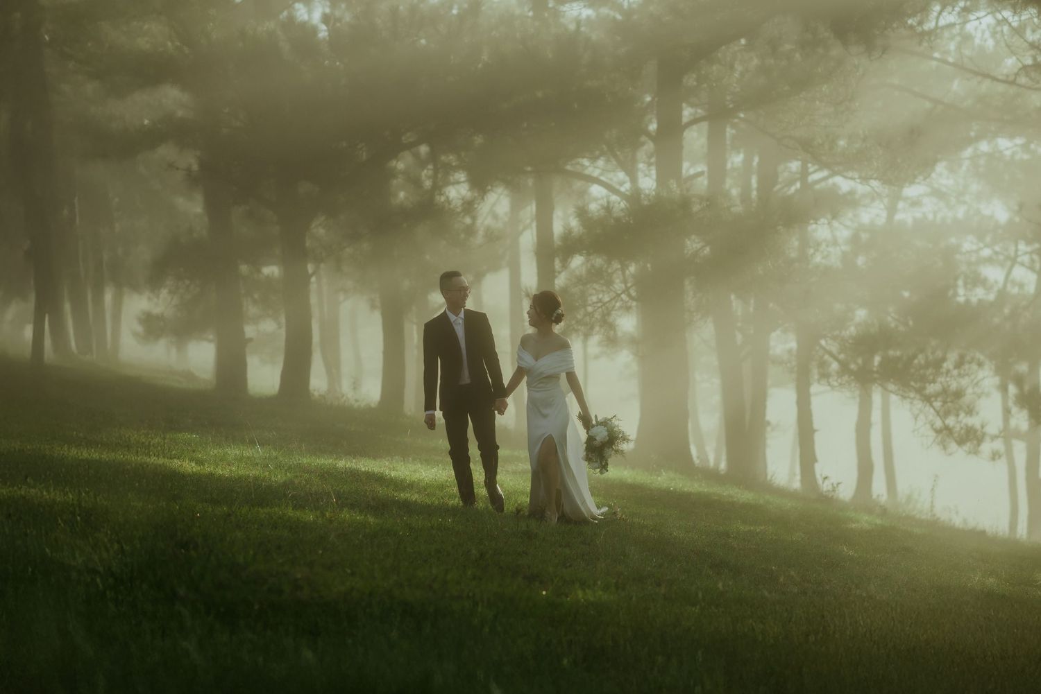 A couple walks hand in hand through a misty forest with soft sunlight filtering through the trees.