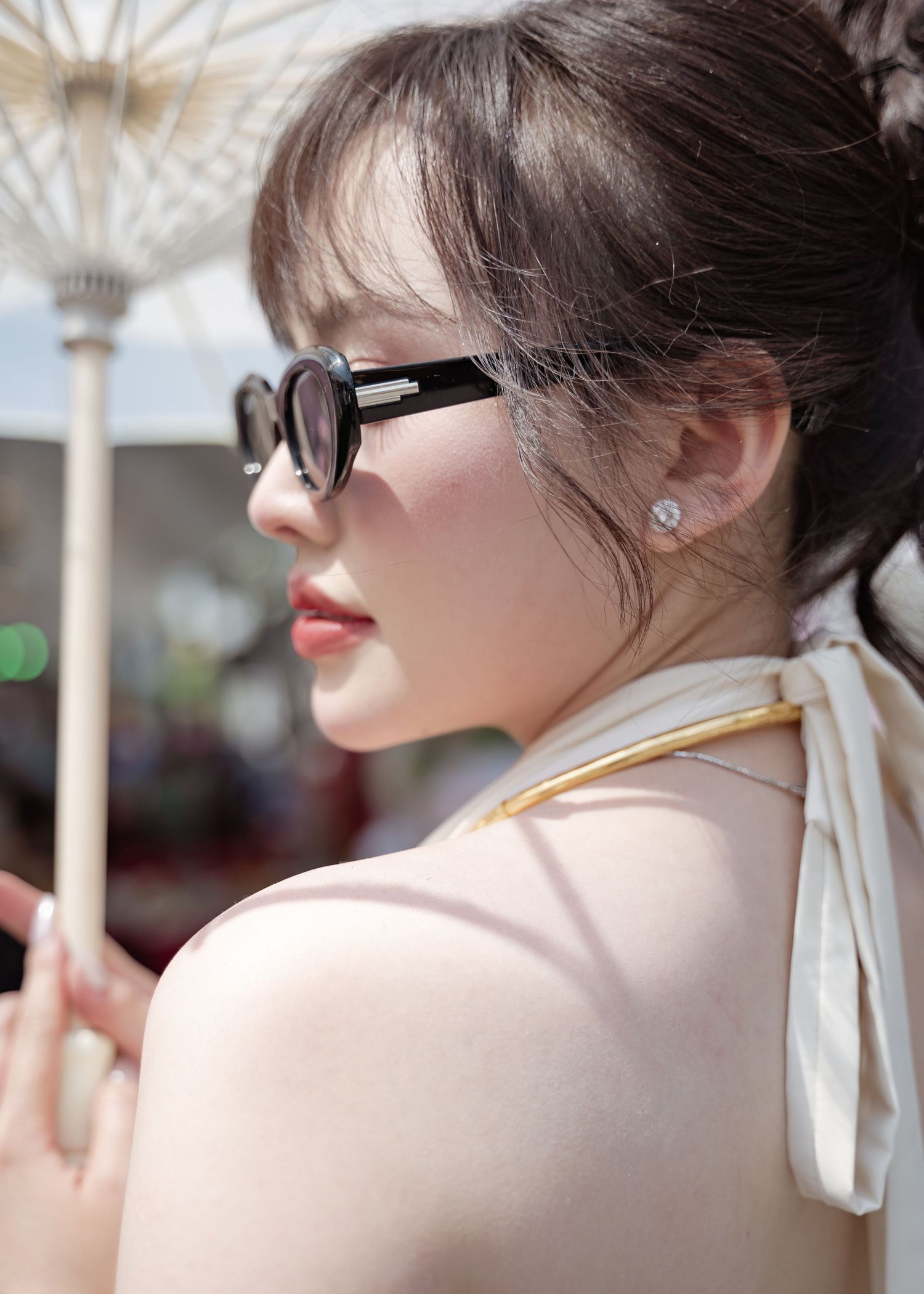 Profile view of someone in sunglasses and a cream colored top with a white ribbon accessory, taken outdoors.