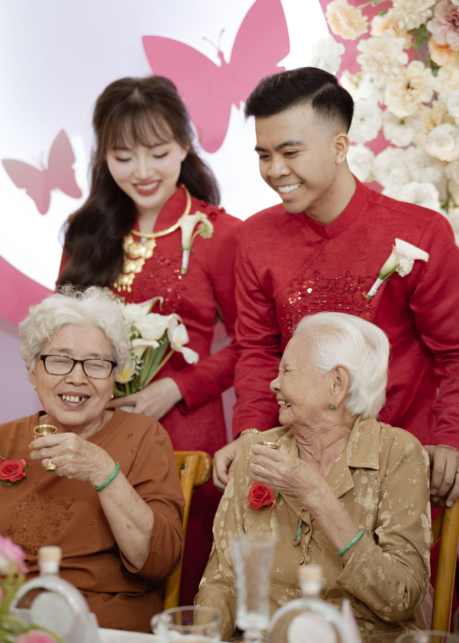 A festive gathering with two couples in traditional red Chinese attire and pink butterflies in the background.
