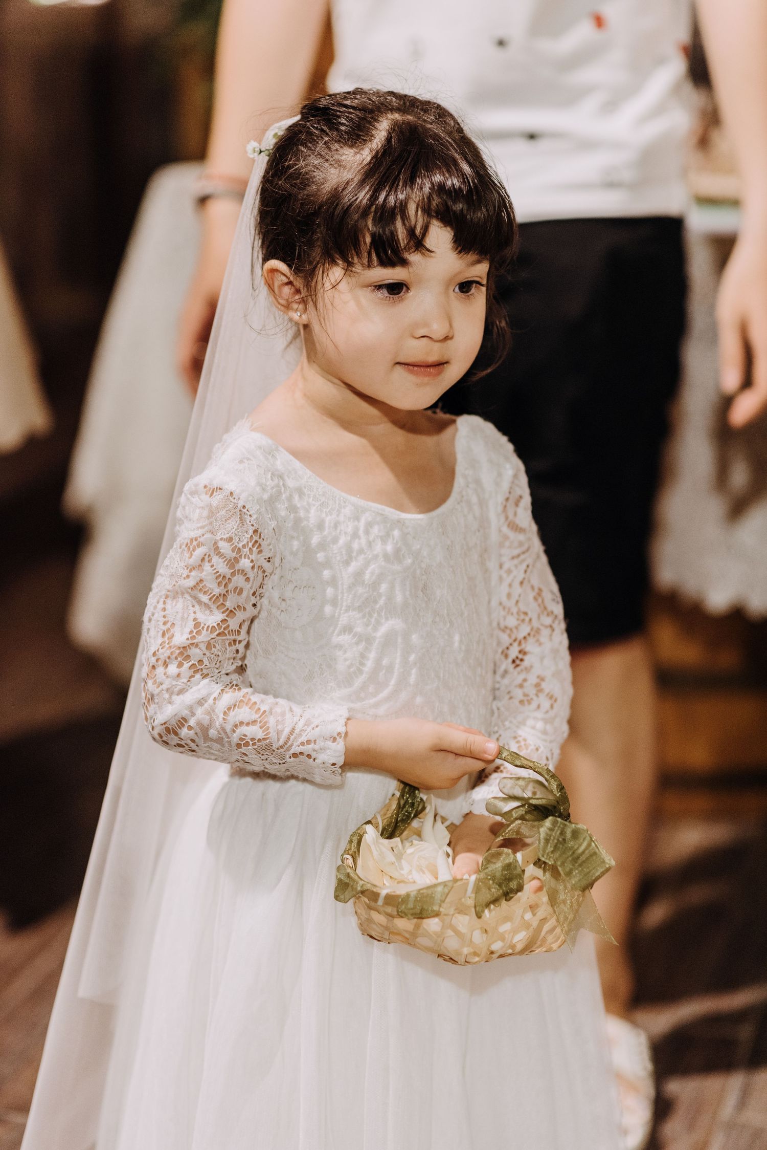A flower girl in a white lace dress holds a small basket at a wedding.