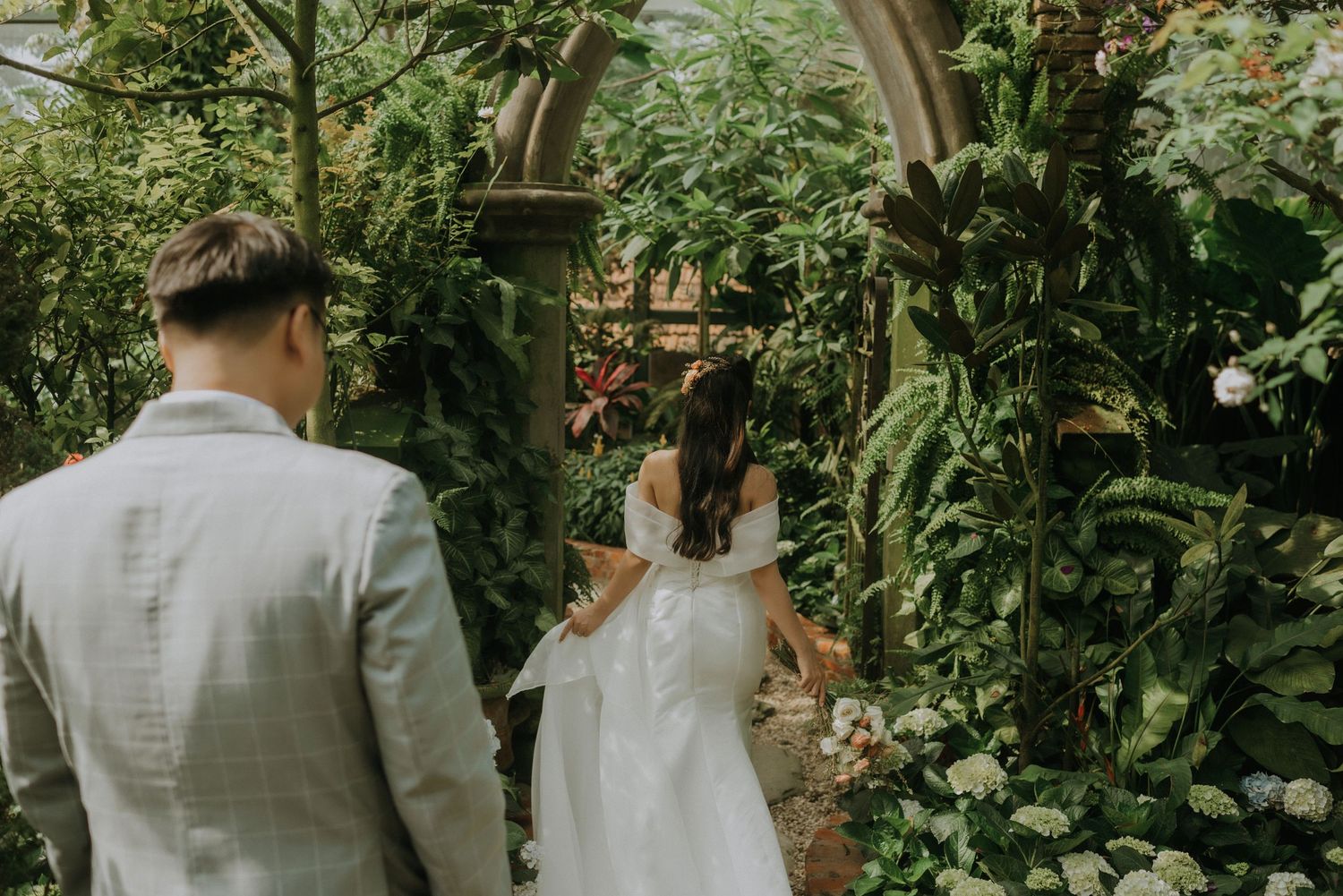In a lush greenhouse garden setting, a couple in wedding attire stands surrounded by verdant plants and flowers.