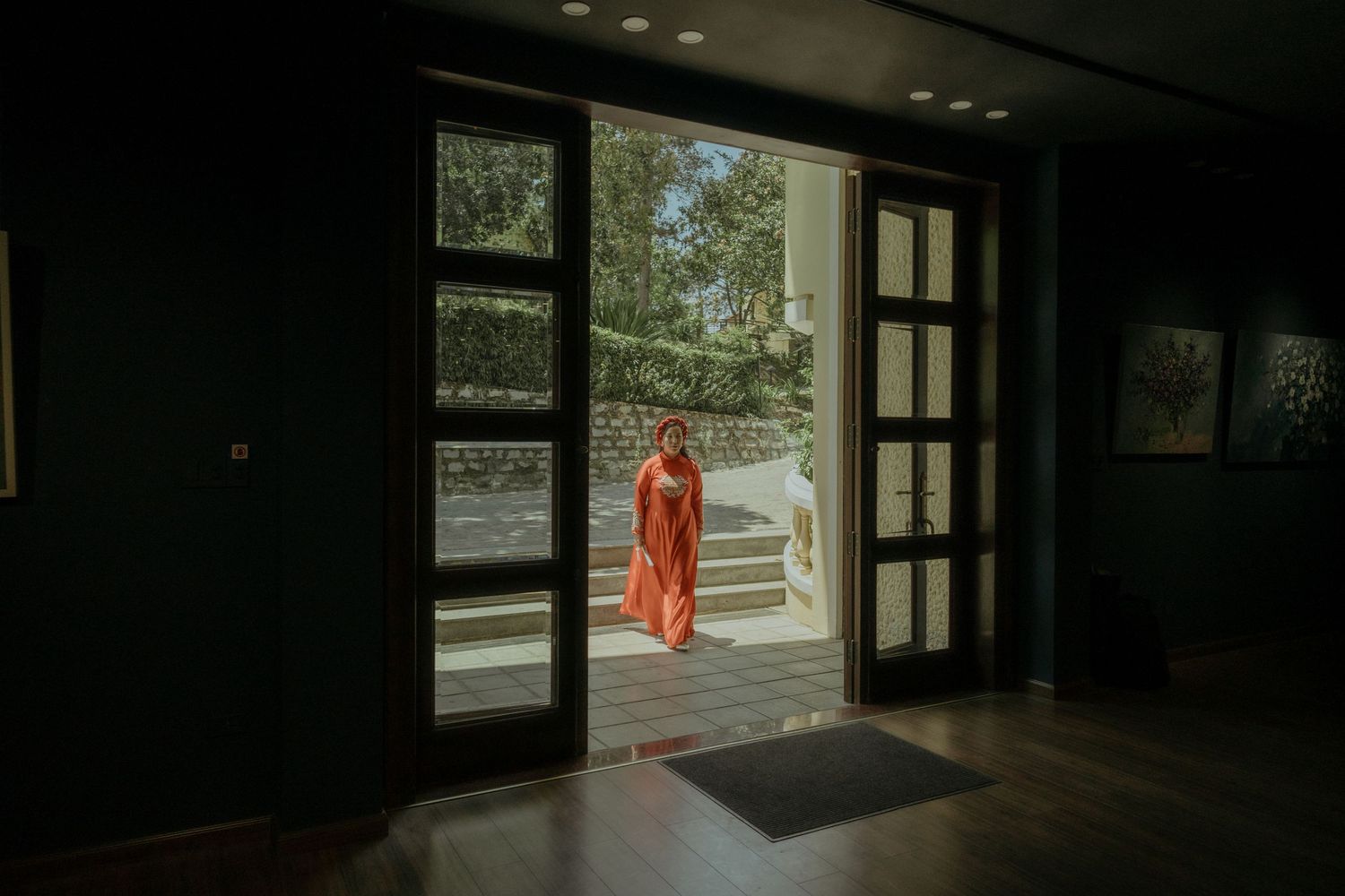 Through a dark doorway, a person in a bright red dress pauses silently on a sunlit exterior patio.