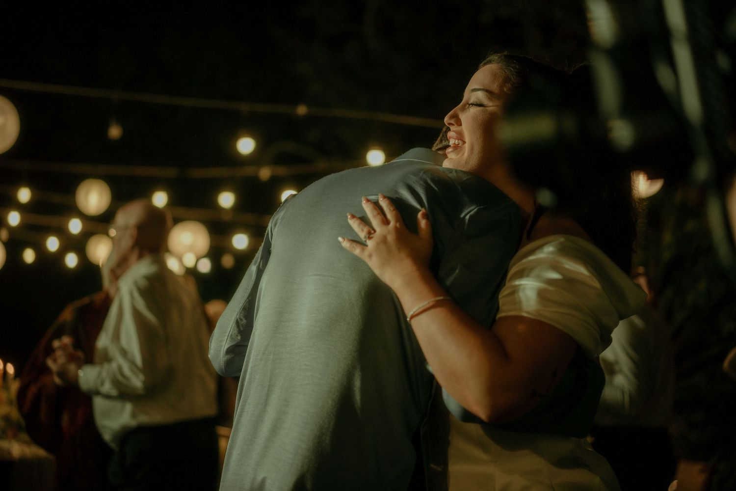 Two people share an emotional embrace during a dimly lit evening celebration with bokeh lights in the background.