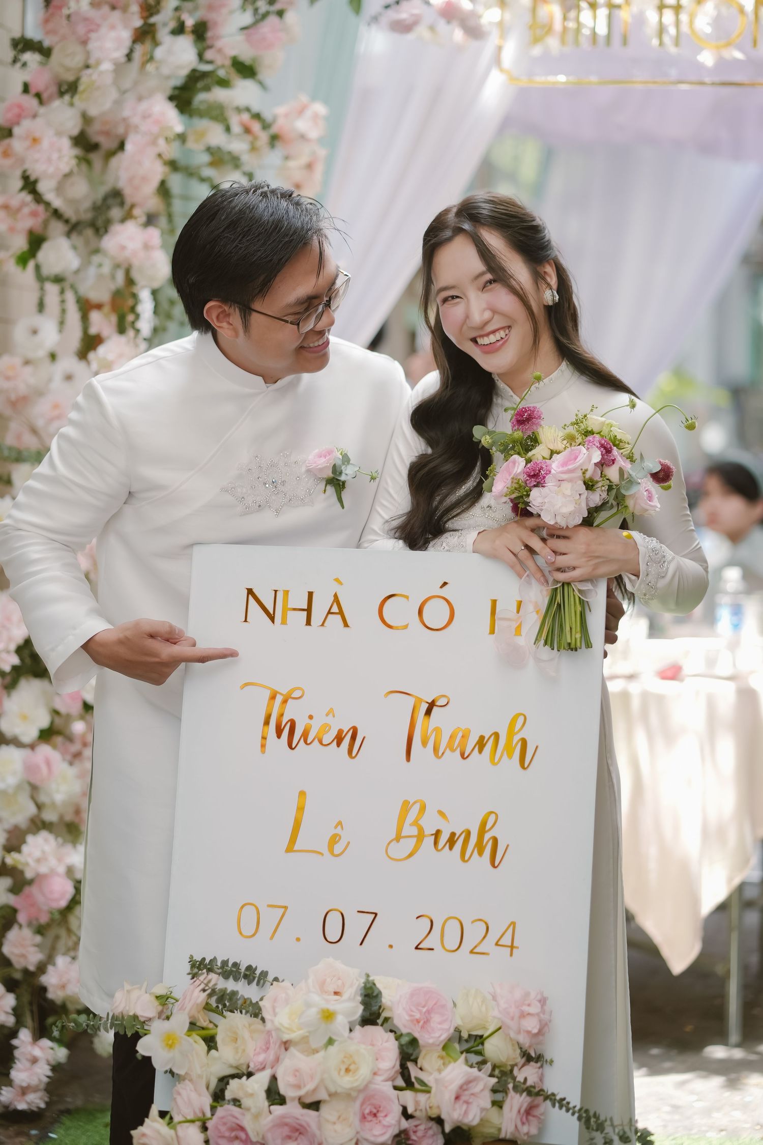 Wedding couple in white attire holding a gold-lettered sign with floral decorations.
