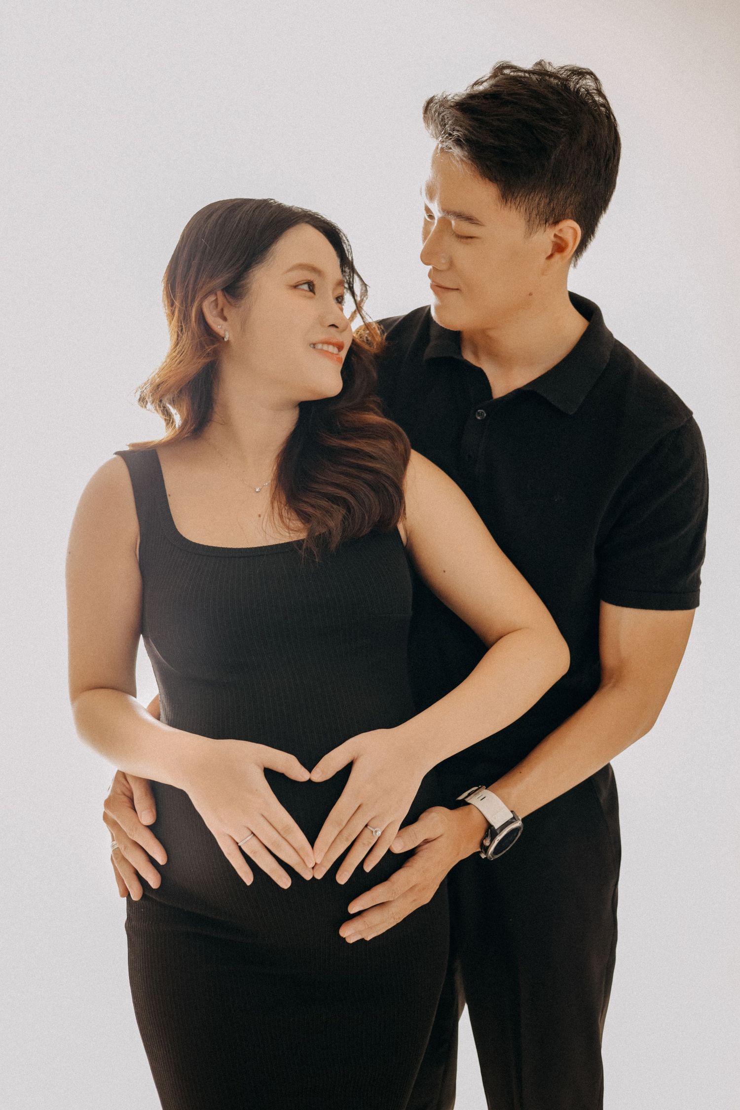 A couple in black outfits forms a heart shape on her pregnant belly in a studio setting.