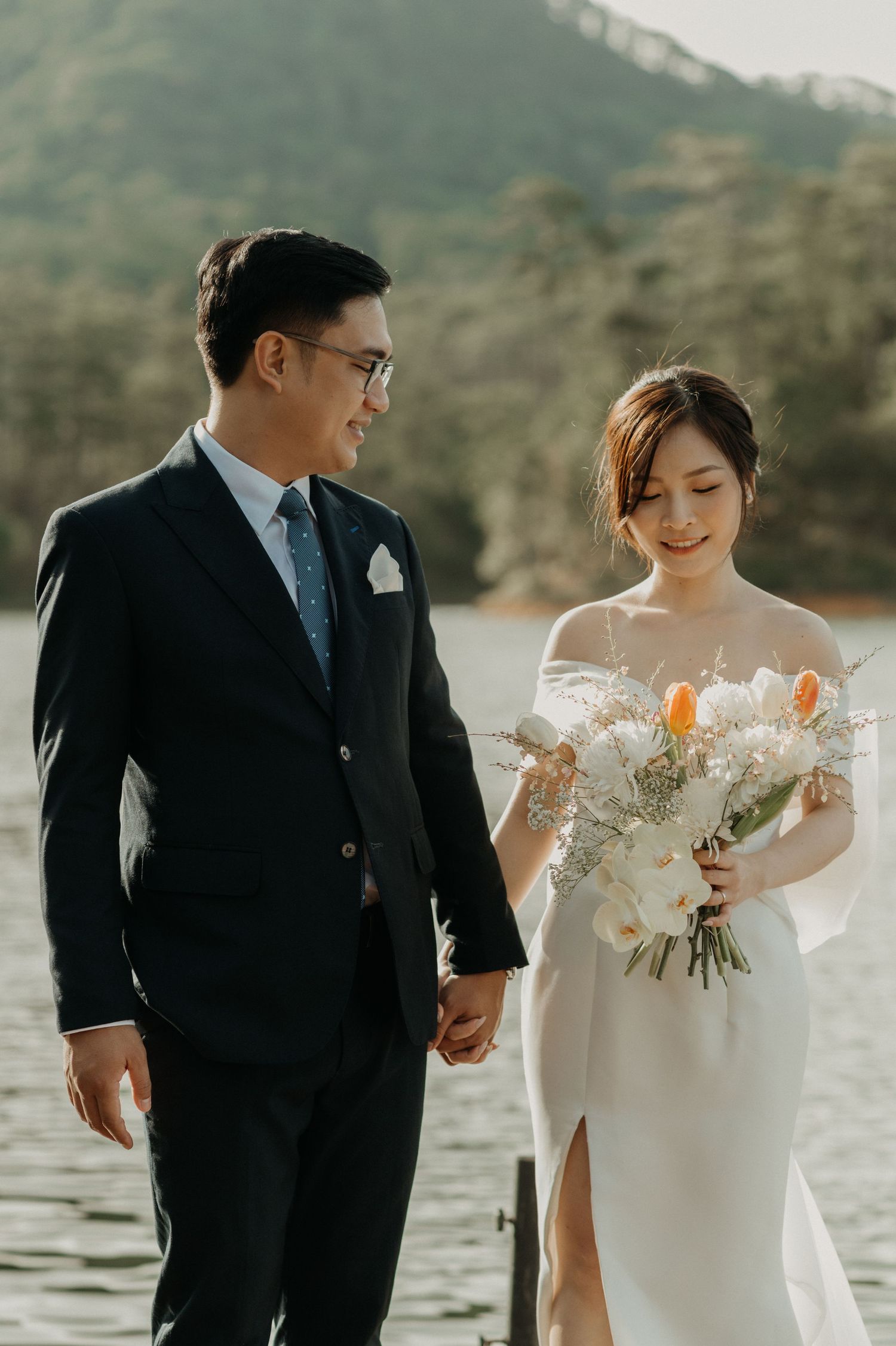 A wedding couple holds hands by a lake wearing elegant attire with the bride carrying a white and orange bouquet.