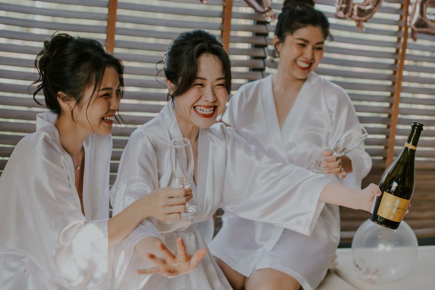 Friends in matching white robes share champagne and laughter during wedding morning preparations.