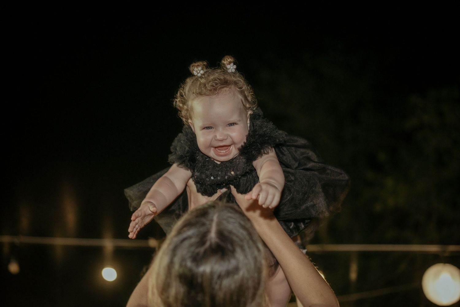 A small child wearing a black dress is lifted up and silhouetted against a dark evening sky with bokeh lights in background.