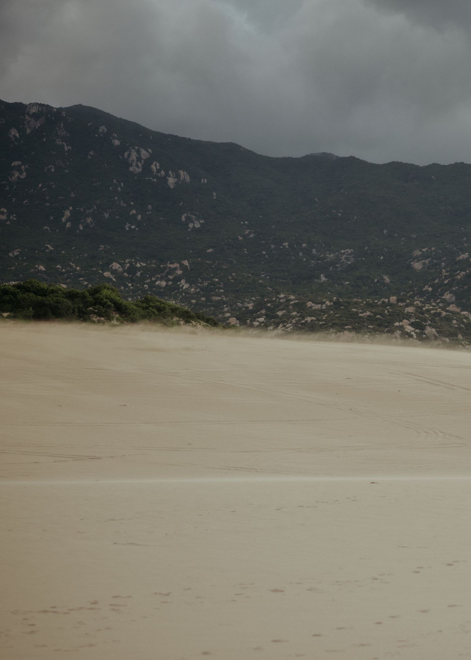 A sandy beach stretches beneath dark mountains and moody storm clouds, creating a dramatic coastal landscape.