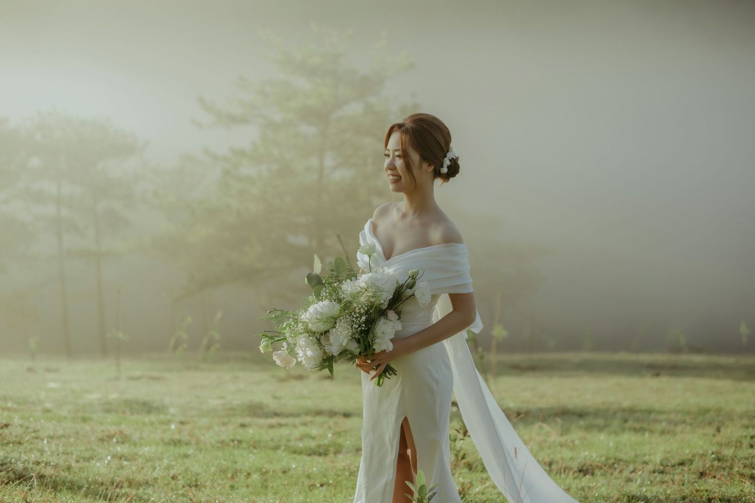 Ethereal wedding scene in misty field with flowing white dress and bouquet of white flowers during golden hour.