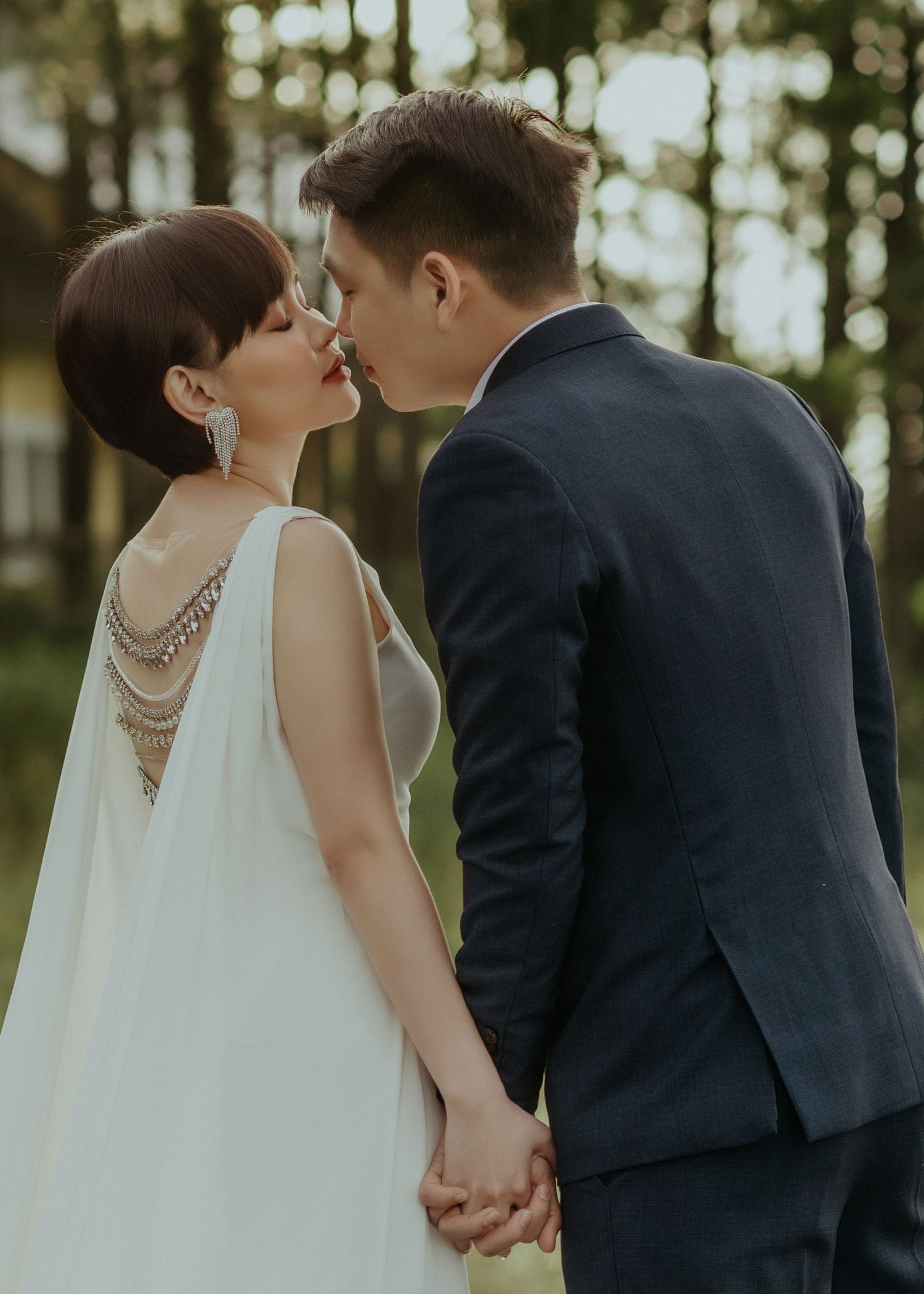 A romantic wedding couple shares an intimate kiss in a forest setting, with the bride wearing a flowing dress with decorative back.