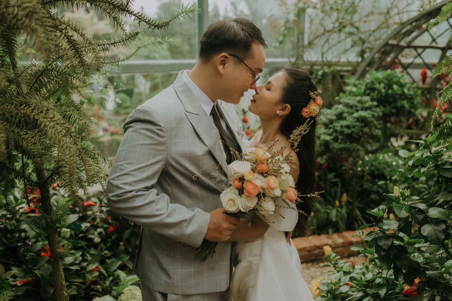 A couple shares a romantic moment in a lush greenhouse garden while holding a peach and coral floral bouquet.