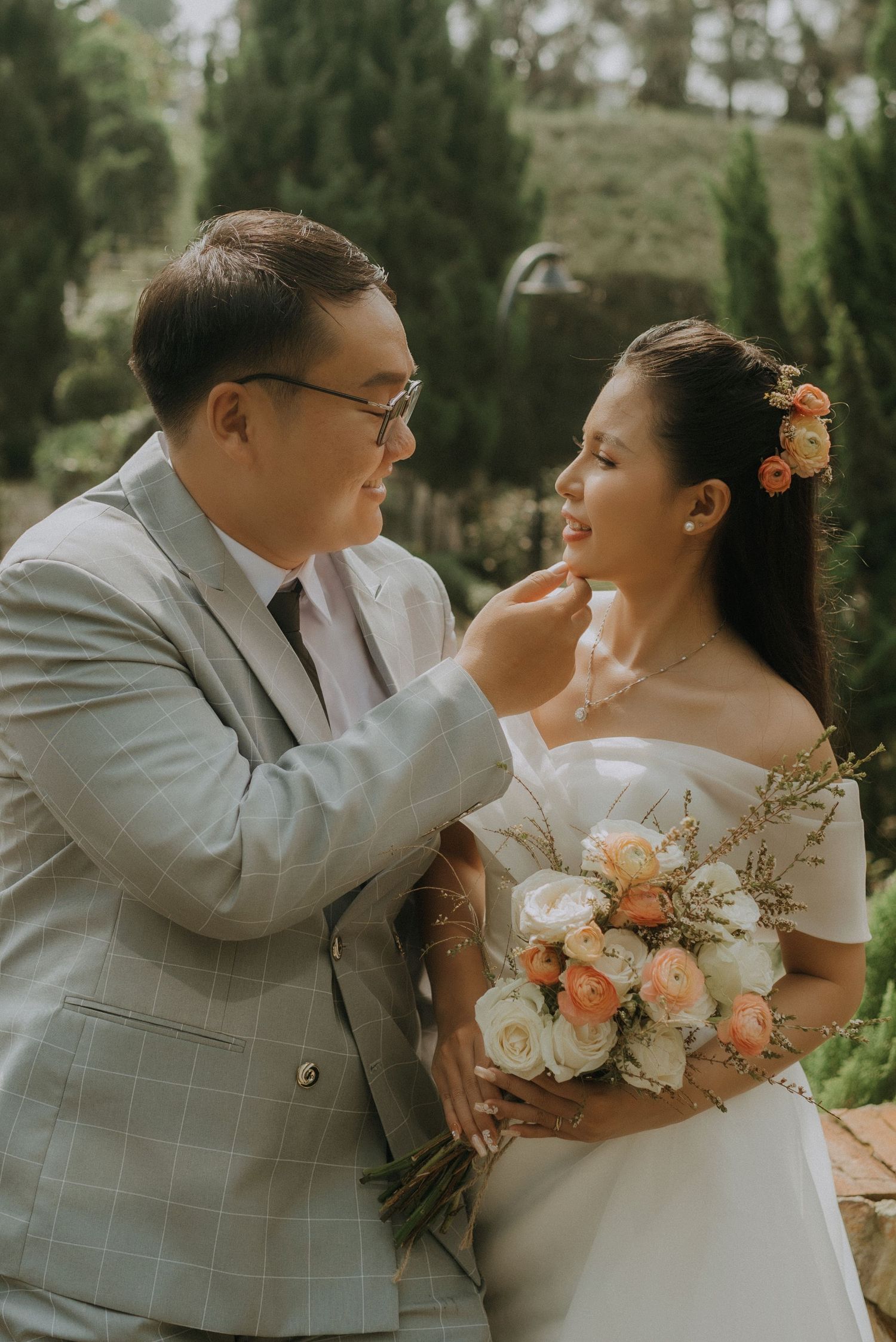 A romantic wedding couple shares an intimate moment in a garden setting with the bride holding a peach and white bouquet.
