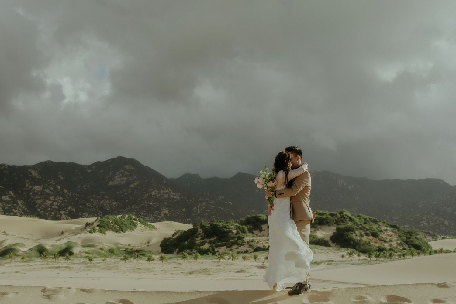 A couple embraces under dramatic storm clouds at a desert landscape with mountains in the background.