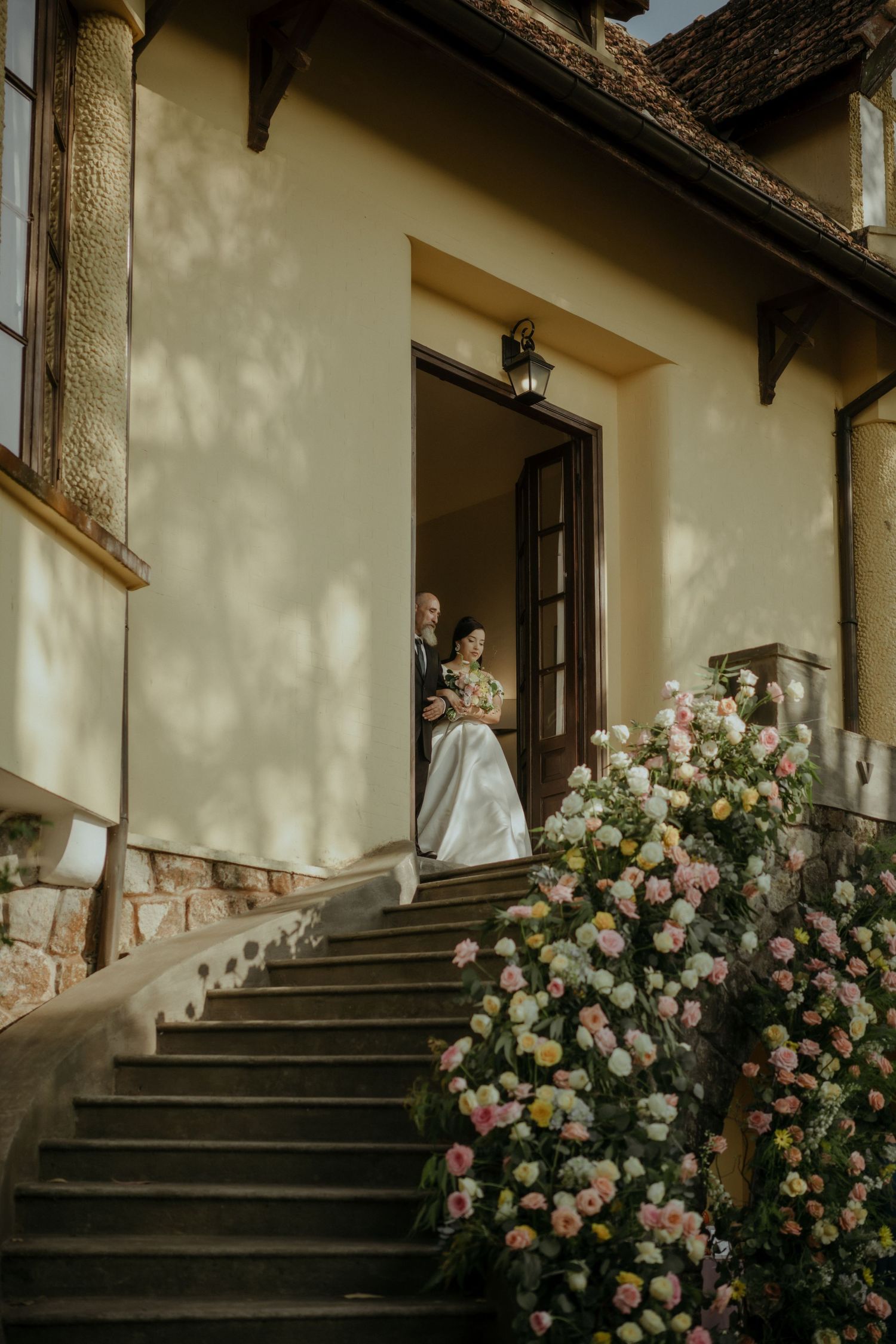 Elegant stone staircase adorned with blooming pink roses leads to manor house entrance in warm afternoon light.