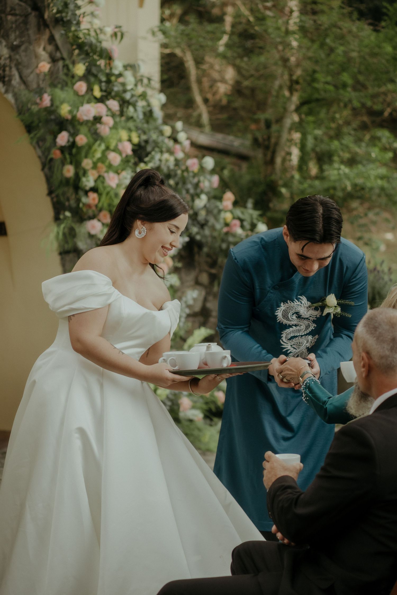 A couple participates in a traditional wedding tea ceremony surrounded by blooming flowers in an outdoor garden setting.