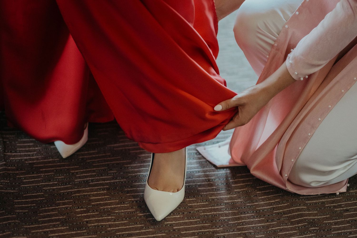 Someone in a red dress being helped with putting on elegant white bridal shoes on a dark wood floor.
